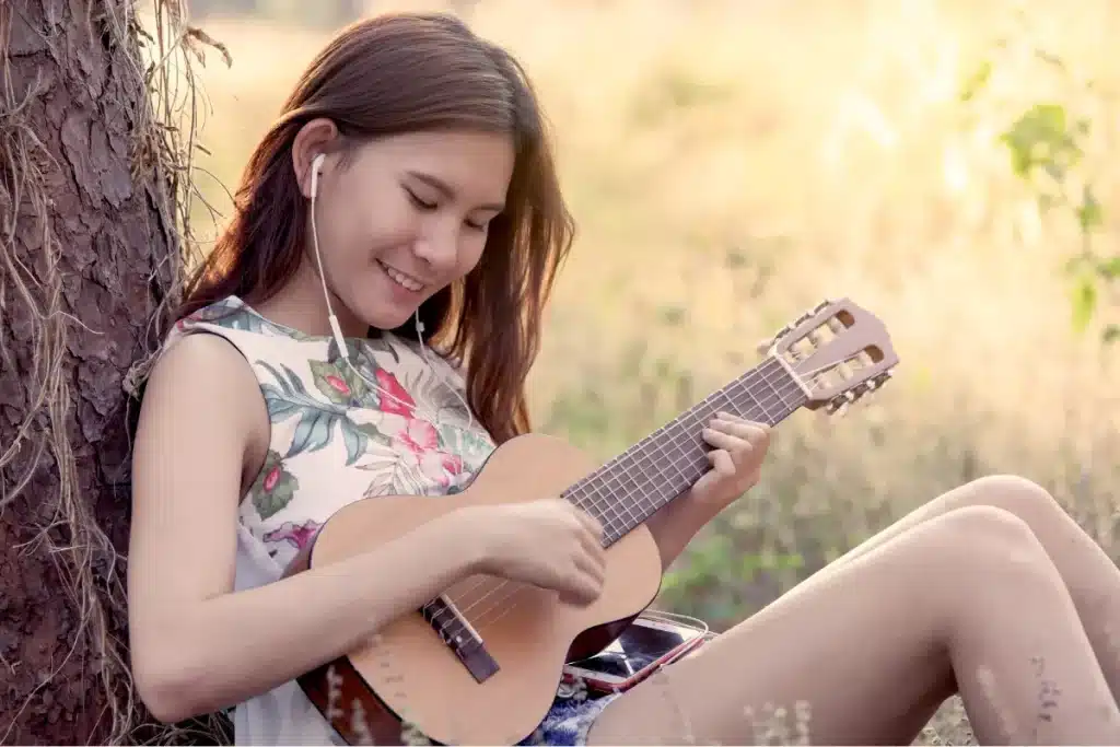 Jeune femme jouant du guitalélé en extérieur. C'est entre le ukulélé et la guitare il y a des différences